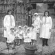 Note Gruppo di bambini con vestitini bianchi e cuffiette assistiti da due donne istitutrici, nel giardino dellasilo, al bordo di una vasca.