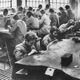 Il reparto bigiottieri. Uomini e donne ai banchi di lavoro. Illuminazione con numerose lampade ad incandescenza.